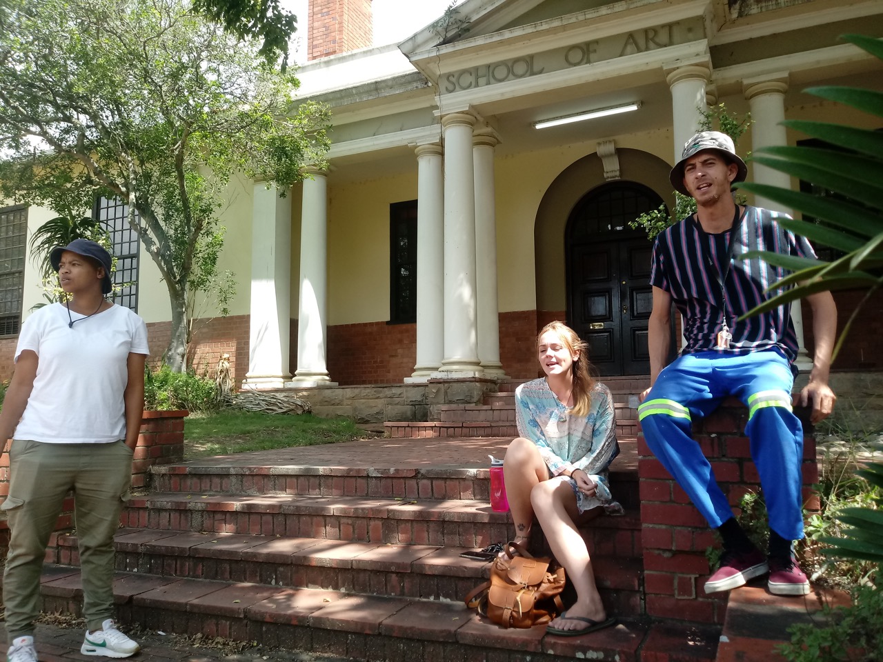 Phila Phaliso and Emily Lacon-Allin sit with Mook Lion in front of the fine art department of Rhodes University. Phila is a master’s student in fine art and the tutor of the minor stream students. Emily is also a master’s student who wishes she could be working on the mural.